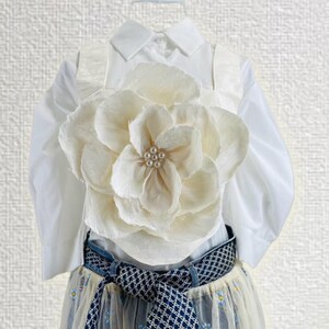 May include: A white button-down shirt with a large cream-colored flower accent. The outfit includes a patterned belt and a sheer skirt with floral details. The overall aesthetic is elegant and feminine.