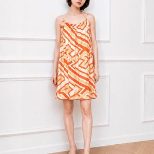 May include: A sleeveless mini dress featuring a vibrant geometric pattern in shades of orange, cream, and tan. The dress has a gathered scoop neckline with thin spaghetti straps and a loose, straight-cut silhouette that falls above the knees.