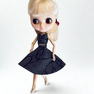 May include: A doll with blonde hair and large brown eyes wearing a black two-piece outfit featuring a textured white pattern. The ensemble consists of a sleeveless crop top and a flared skirt. The doll is wearing light-colored flat shoes.