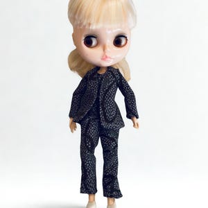 May include: A doll with blonde hair and large brown eyes wearing a black blazer and matching trousers featuring a white geometric pattern. The doll has a pale complexion, pink lips, and is wearing simple light-colored shoes on its feet.