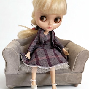 May include: A doll with blonde hair and brown eyes sits on a small gray velvet sofa. The doll wears a dark purple and black plaid dress with a matching jacket and a white tulle underskirt. The doll also wears simple white shoes with ankle straps.