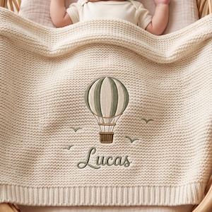 May include: A cream-colored knitted baby blanket featuring an embroidered sage green hot air balloon design with small birds and the name Lucas stitched in a cursive font. The blanket has a textured waffle knit pattern and a ribbed border edge.