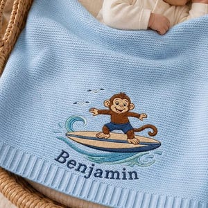 May include: A light blue knitted baby blanket featuring an embroidered graphic of a brown monkey surfing on a blue and tan surfboard. Below the graphic, the name Benjamin is embroidered in dark blue thread. The blanket has a ribbed border.