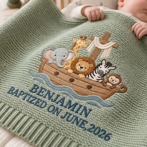 May include: A sage green knitted baby blanket featuring an embroidered brown wooden ark carrying a giraffe, elephant, lion, zebra, and monkey. Below the ark, the text reads BENJAMIN BAPTIZED ON JUNE, 2026 in dark blue embroidered lettering.
