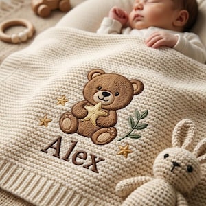 Könnte beinhalten: Eine cremefarbene Strickdecke für Babys mit einem gestickten braunen Teddybären, der einen gelben Stern hält, umgeben von kleinen goldenen Sternen und einem grünen Blattzweig. Der Name Alex ist in braunem Garn unter dem Bärenmotiv eingestickt.