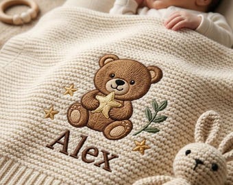 May include: A cream-colored knitted baby blanket featuring an embroidered brown teddy bear holding a yellow star, surrounded by small gold stars and a green leafy sprig. The name Alex is embroidered in brown thread below the bear design.