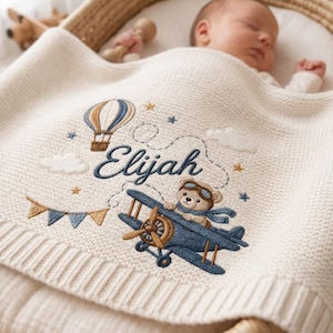 May include: A cream-colored knit baby blanket featuring an embroidered design of a teddy bear in a blue biplane, a hot air balloon, clouds, stars, and bunting. The name Elijah is embroidered in dark blue script in the center of the blanket.
