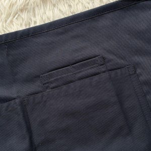 Op de afbeelding: Close-up van een marineblauwe stof met een zakdetail. De zak is rechthoekig en op de hoofdstof gestikt. De stof heeft een gladde textuur en is effen donkerblauw.