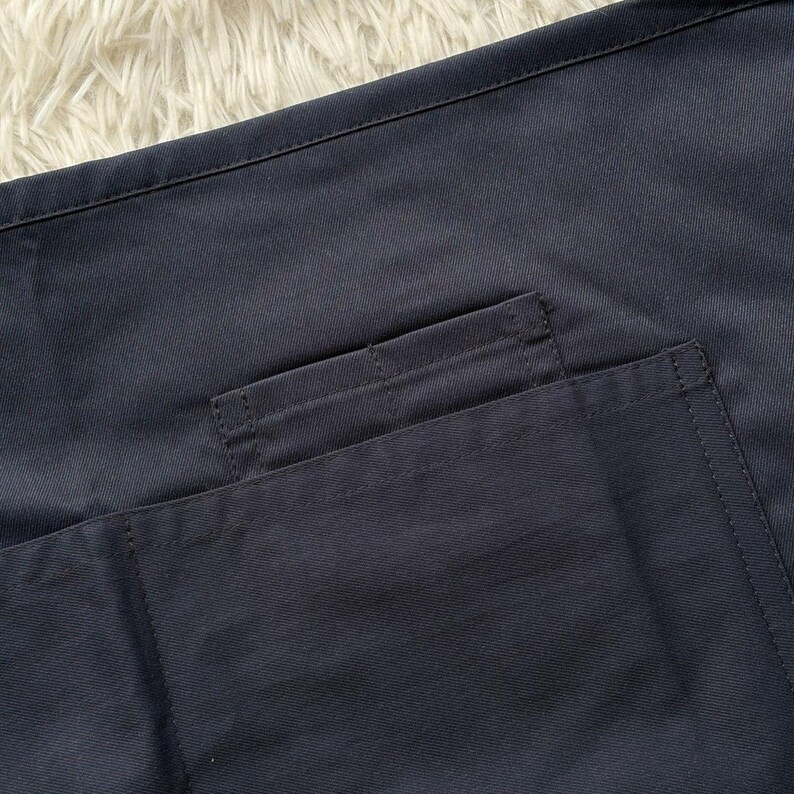 Op de afbeelding: Close-up van een marineblauwe stof met een zakdetail. De zak is rechthoekig en op de hoofdstof gestikt. De stof heeft een gladde textuur en is effen donkerblauw.