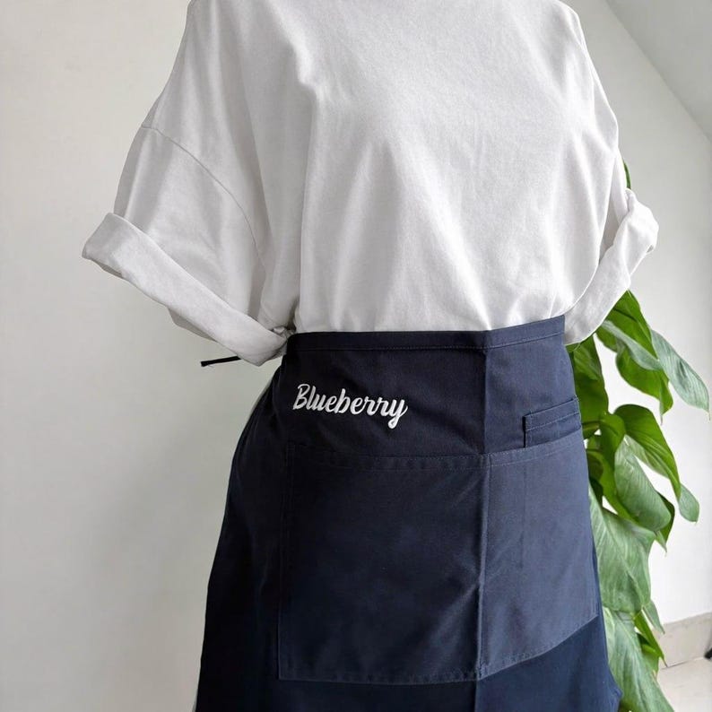 Op de afbeelding: Een marineblauwe schort met twee voorzakken en het woord "Blueberry" geborduurd in witte letters. De schort wordt gedragen over een wit t-shirt met opgerolde mouwen. De schort is gemaakt van een duurzame stof.