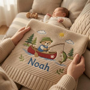 May include: A beige knit blanket featuring an embroidered scene of a bear in a red canoe fishing for a green fish. The design includes a yellow sun, blue clouds, green pine trees, and the name Noah stitched in blue thread at the bottom centre.