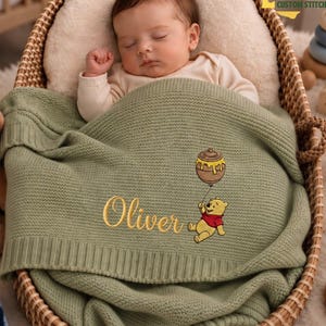 Pode incluir: Um bebê dormindo está aninhado em uma cesta de vime, coberto por um cobertor de malha verde sálvia. O cobertor apresenta o nome "Oliver" bordado em dourado, junto com um desenho de Ursinho Pooh segurando um balão de pote de mel.