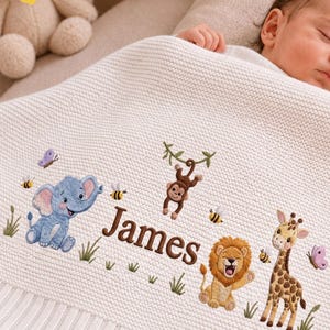 Könnte beinhalten: Eine cremefarbene Strickdecke für Babys mit gestickten Dschungeltieren, darunter ein blauer Elefant, ein brauner Affe, ein Löwe und eine Giraffe, sowie Bienen und Schmetterlinge. Der Name James ist mittig in brauner Schrift eingestickt.
