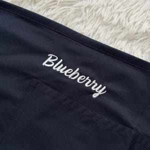 Op de afbeelding: Close-up van een marineblauwe schort met een zak. Het woord "Blueberry" is in witte cursieve letters geborduurd over de bovenkant van de schort. De schort is gemaakt van een gladde, donkere stof.