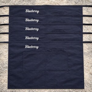 Op de afbeelding: Vijf marineblauwe schorten met het woord "Blueberry" in witte letters geborduurd. Elk schort heeft een grote voorzak en lange zwarte banden. De schorten worden gepresenteerd op een lichtgekleurd, getextureerd oppervlak.