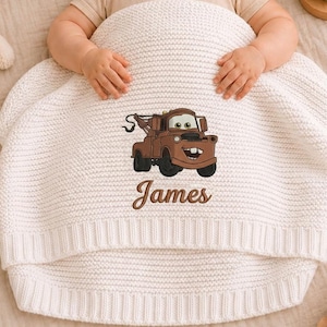 May include: A cream-colored knitted baby blanket featuring an embroidered brown cartoon tow truck character. The name "James" is embroidered below the truck. The blanket is wrapped around a baby.