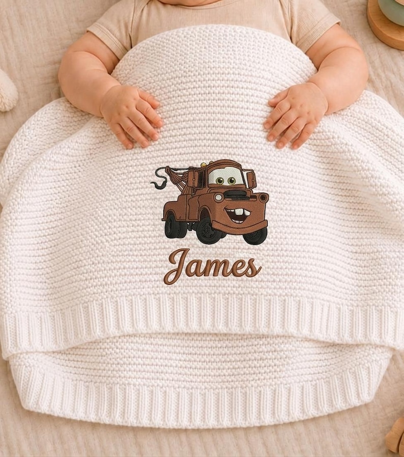 May include: A cream-colored knitted baby blanket featuring an embroidered brown cartoon tow truck character. The name "James" is embroidered below the truck. The blanket is wrapped around a baby.