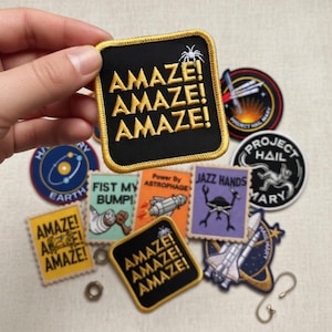 Może przedstawiać: Kolekcja haftowanych naszywek z różnymi wzorami i tekstami. Centralna naszywka, trzymana w dłoni, jest czarna ze złotym napisem "AMAZE!" powtórzonym trzy razy. Inne naszywki przedstawiają grafikę o tematyce kosmicznej i frazy takie jak "JAZZ HANDS" i "PROJECT HAIL MARY".