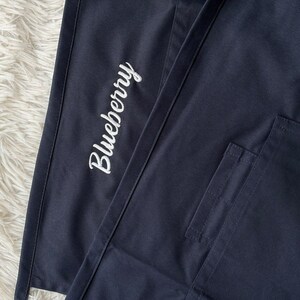 Op de afbeelding: Close-up van marineblauwe stof met het woord "Blueberry" in witte letters geborduurd. Een zak is zichtbaar aan de rechterkant van de afbeelding. De stof lijkt deel uit te maken van een schort of werkkleding.
