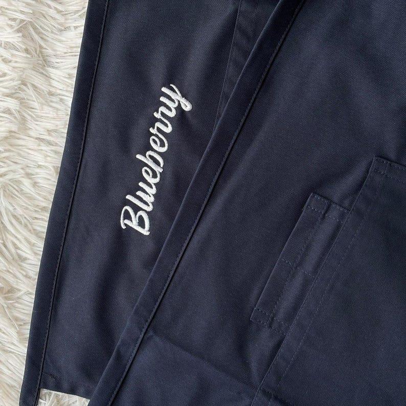 Op de afbeelding: Close-up van marineblauwe stof met het woord "Blueberry" in witte letters geborduurd. Een zak is zichtbaar aan de rechterkant van de afbeelding. De stof lijkt deel uit te maken van een schort of werkkleding.