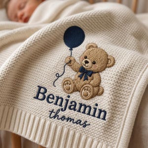 May include: A cream-colored knitted baby blanket featuring an embroidered brown teddy bear holding a dark blue balloon. The bear wears a dark blue bow tie. The name Benjamin is embroidered in dark blue block letters with thomas below it.