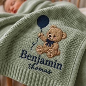 May include: A sage green knitted baby blanket featuring an embroidered brown teddy bear holding a dark blue balloon. The name Benjamin is embroidered in dark blue capital letters above the name thomas in dark blue lowercase script lettering.