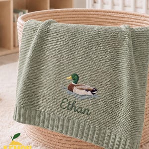 Könnte beinhalten: Eine salbeigrüne Strickdecke mit einer Stickerei einer Stockente, die auf dem Wasser schwimmt, und dem Namen Ethan in dunkelgrüner Schreibschrift darunter. Die Decke hat einen gerippten Rand an der Unterseite und ein strukturiertes Strickmuster.