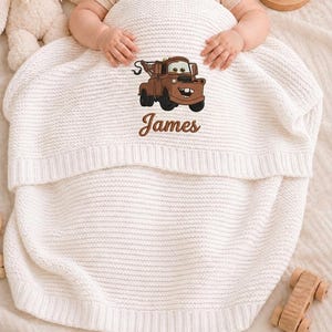 May include: A white knitted baby blanket featuring an embroidered cartoon tow truck design in brown and black. The name "James" is embroidered below the truck. The blanket is folded over a baby.