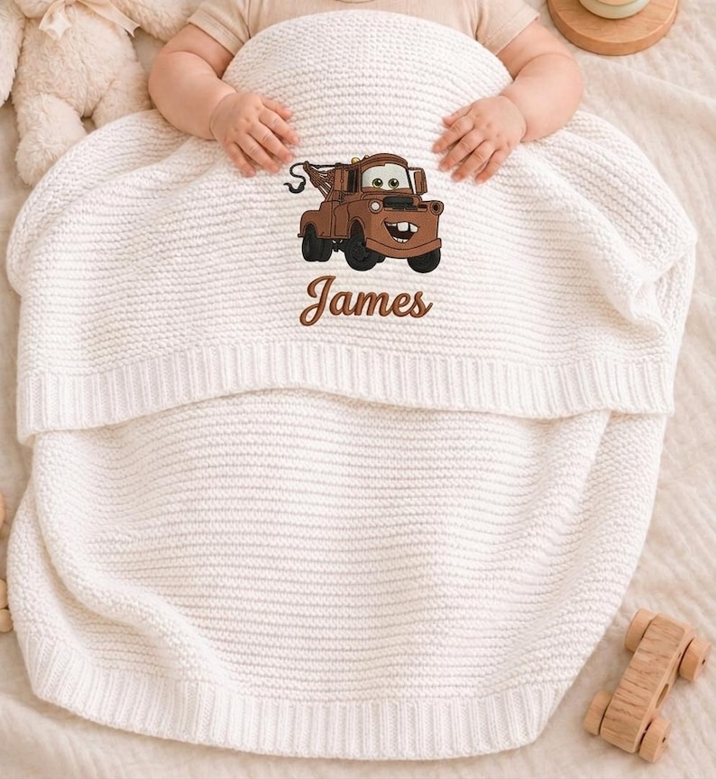 May include: A white knitted baby blanket featuring an embroidered cartoon tow truck design in brown and black. The name "James" is embroidered below the truck. The blanket is folded over a baby.