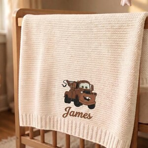 May include: Cream-colored knitted baby blanket draped over a wooden crib. The blanket features an embroidered image of a brown cartoon tow truck and the name "James" in a script font. The blanket has a ribbed border.