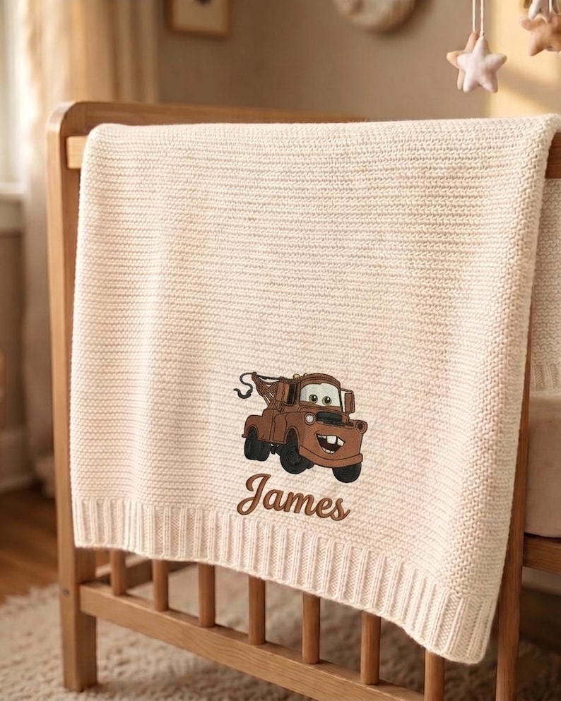May include: Cream-colored knitted baby blanket draped over a wooden crib. The blanket features an embroidered image of a brown cartoon tow truck and the name "James" in a script font. The blanket has a ribbed border.