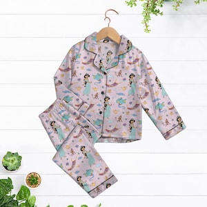 Op de afbeelding: Roze pyjamaset met een herhaald patroon van een prinses, een magische lamp en andere personages. De top met lange mouwen en de broek hebben een zwarte bies. De set hangt aan een houten hanger tegen een witte achtergrond.