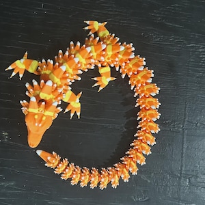 May include: A whimsical, orange and yellow dragon sculpture, crafted to resemble candy corn. The dragon features a long, curved body with detailed scales and pointed limbs. The sculpture is set against a dark background, highlighting its vibrant colors and unique design.