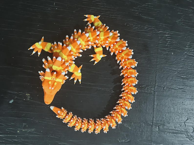 May include: A whimsical, orange and yellow dragon sculpture, crafted to resemble candy corn. The dragon features a long, curved body with detailed scales and pointed limbs. The sculpture is set against a dark background, highlighting its vibrant colors and unique design.