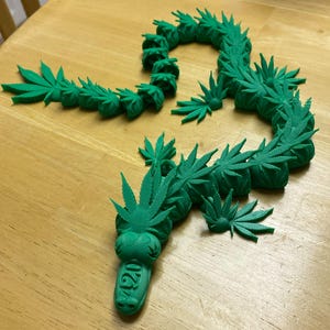May include: A green, 3D-printed sculpture of a dragon, crafted from cannabis leaves. The dragon's head features the number "420" on its snout. The sculpture is displayed on a wooden surface.