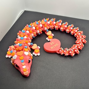 May include: A pink dragon figurine decorated with colorful heart-shaped accents. The dragon has a red heart-shaped charm that says "BE MINE". The figurine is on a black surface.