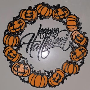 May include: A circular wall decoration featuring a ring of orange jack-o'-lanterns and plain pumpkins connected by black vine-like structures. In the center, the text "happy Halloween" is written in a black, cursive, stylized script font.