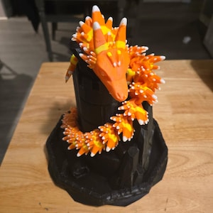 May include: A 3D-printed dragon sculpture with a black base and body, adorned with orange, yellow, and white candy corn scales. The dragon's head is orange, with candy corn accents. The sculpture is on a wooden surface.
