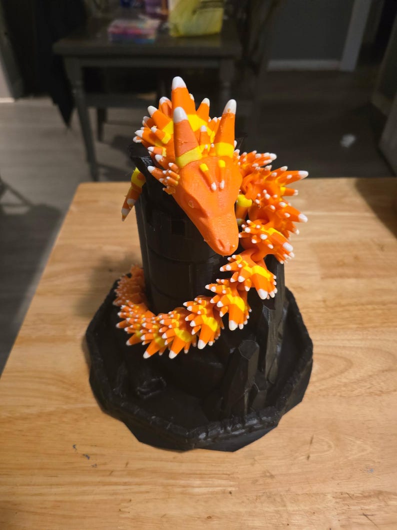 May include: A 3D-printed dragon sculpture with a black base and body, adorned with orange, yellow, and white candy corn scales. The dragon's head is orange, with candy corn accents. The sculpture is on a wooden surface.