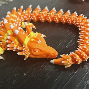 May include: A 3D-printed orange dragon toy with candy corn-like details. The dragon has a detailed head, body, and tail, with white, yellow, and orange segments. The toy is flexible and can be posed.