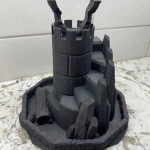 May include: A black 3D-printed castle model with a cylindrical tower, steps, and jagged rock formations. The model sits on a circular base with a textured surface. The castle has a detailed, textured appearance.