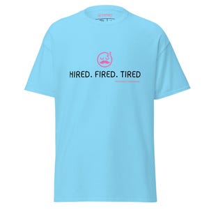 May include: A light blue t-shirt with the text "HIRED. FIRED. TIRED" in black, and a pink sleeping face with a mustache. The shirt is made of a soft material and has a crew neck.