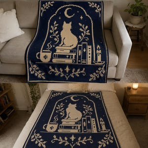 May include: A dark blue and cream knit throw blanket featuring a pixelated design of a cat sitting on a stack of books, a steaming mug, a candle, and a crescent moon framed by an archway with botanical accents and stars throughout the pattern.