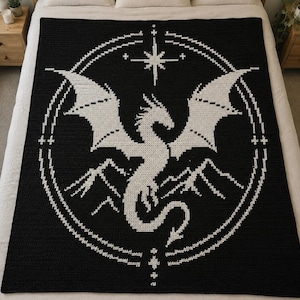 May include: A black and white crochet blanket featuring a large central dragon silhouette surrounded by a circular frame. The design includes a stylized star at the top and mountain peaks at the bottom, all rendered in a pixelated stitch pattern.