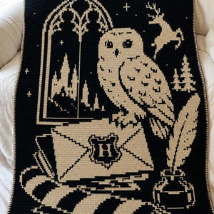 May include: A rectangular crochet blanket featuring a black and cream colour scheme. The design depicts a snowy owl perched on an envelope with an H crest, a quill in an inkwell, a castle silhouette through a window, and a stag silhouette.