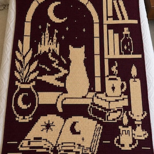 May include: A rectangular crochet blanket in deep burgundy with a cream-colored tapestry design. The pattern features a cat sitting on a windowsill, a castle, a crescent moon, stars, a bookshelf, a potted plant, an open book, and candles.