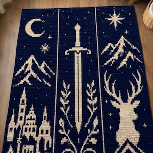 May include: A rectangular crochet blanket in deep navy blue with cream-colored patterns. The design features a central sword flanked by foliage, a crescent moon, stars, mountain ranges, a detailed castle silhouette, and a large stag head.