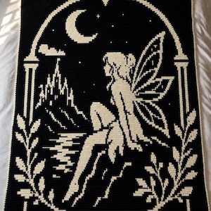 Puede incluir: Una manta de ganchillo en negro y crema que muestra a un hada sentada dentro de un marco arqueado. El diseño incluye una luna creciente, estrellas, la silueta de un castillo y follaje. El hada tiene alas y está junto al agua.