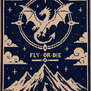 May include: A rectangular dark blue and cream crochet blanket featuring a central dragon inside a circular frame, with the text FLY OR DIE below it. The design includes mountain peaks, a castle at the base, and scattered clouds and stars.