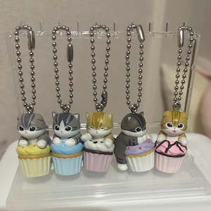 以下が含まれることがあります： 5匹の猫の形をしたキーホルダー。それぞれがカップケーキの上にいます。猫はグレー、オレンジ、茶色など様々な色と模様があります。カップケーキは黄色、青、ピンク、紫、ピンクで、白いフロスティングが施されています。各キーホルダーにはシルバーのボールチェーンが付いています。
